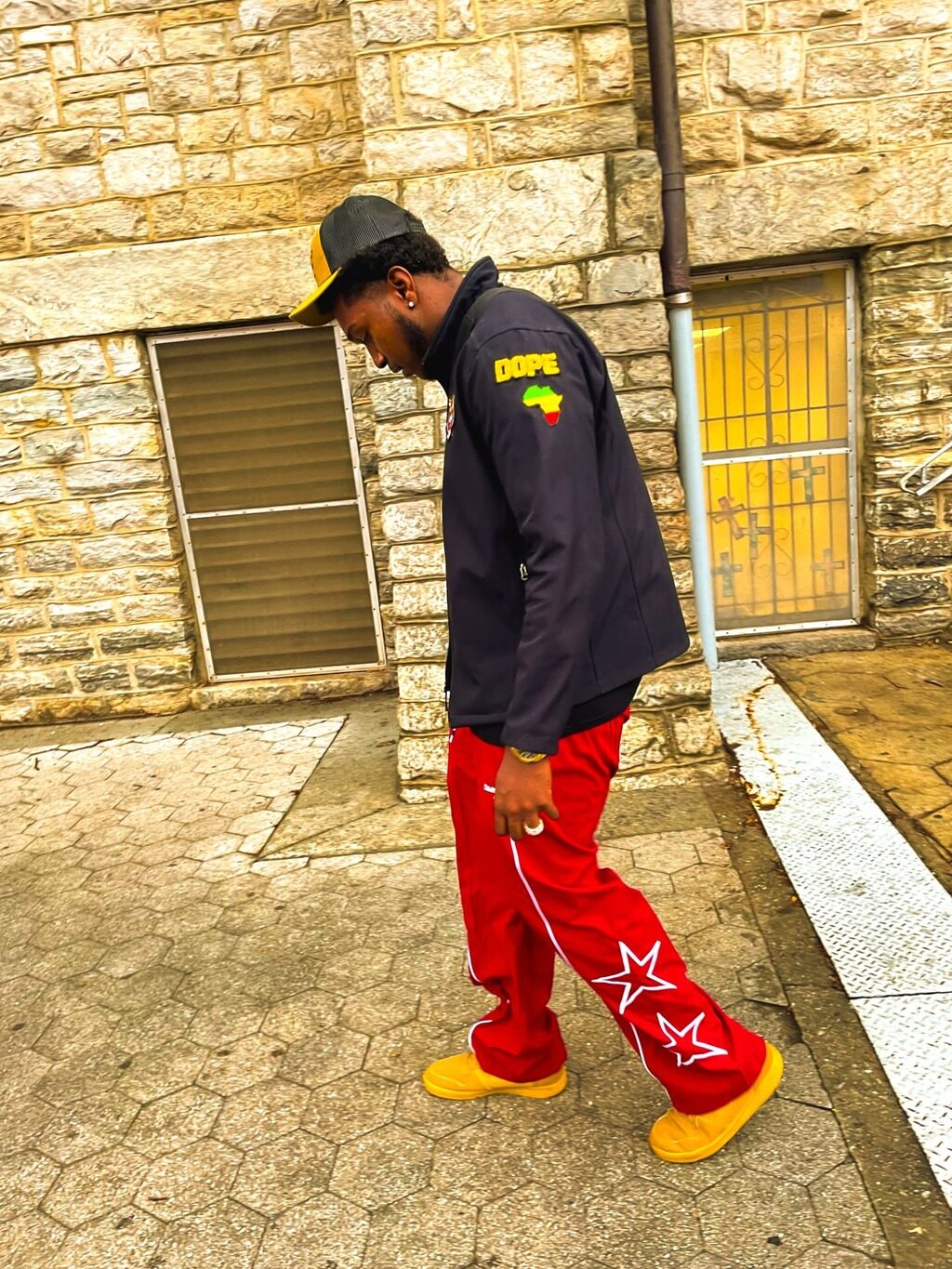 KAPOLLO in profile mid-step in a black DOPE jacket, red star track pants and yellow sneakers against a stone wall.