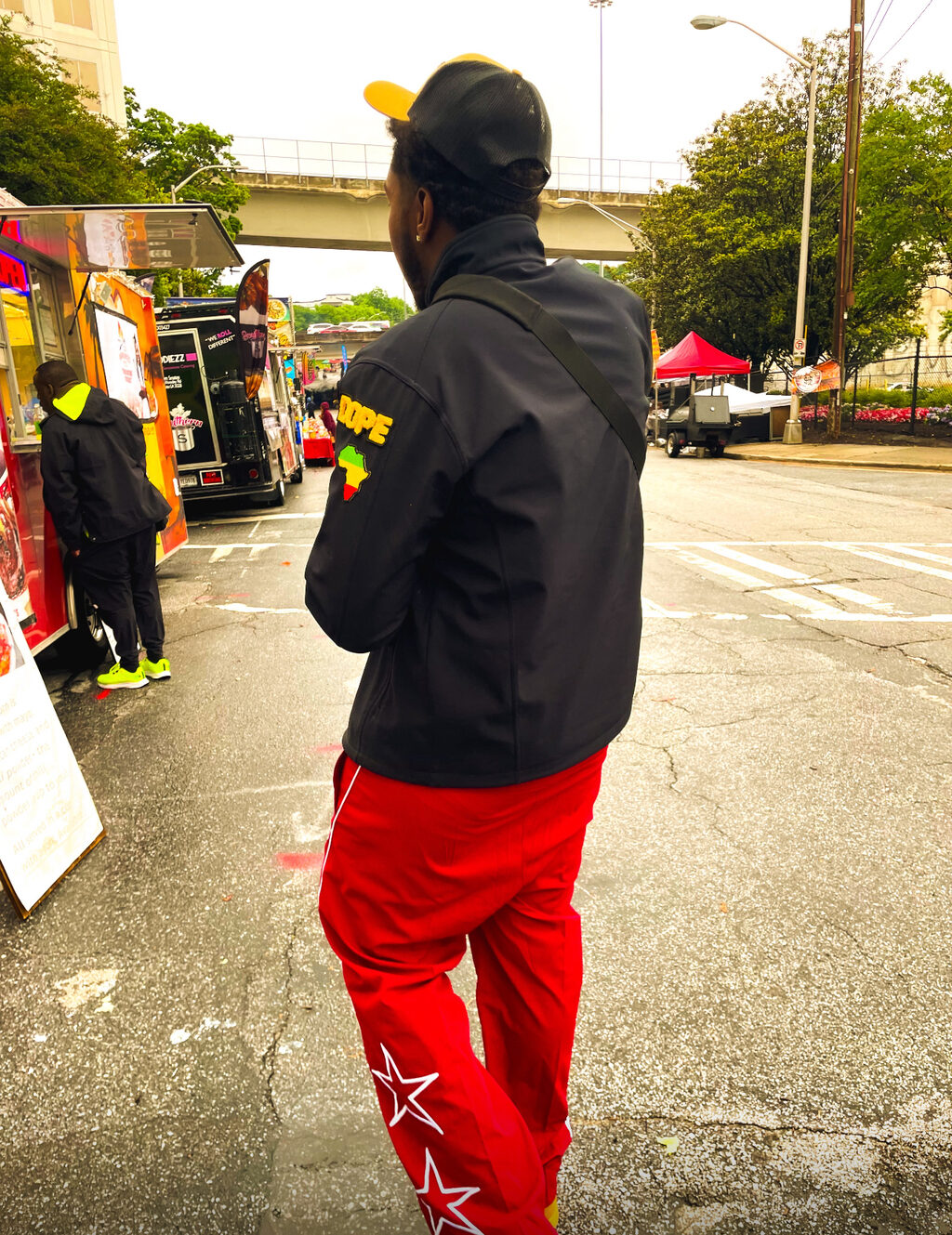 KAPOLLO from behind in a black DOPE jacket and red star track pants, walking past food trucks under an Atlanta overpass on a wet grey day.