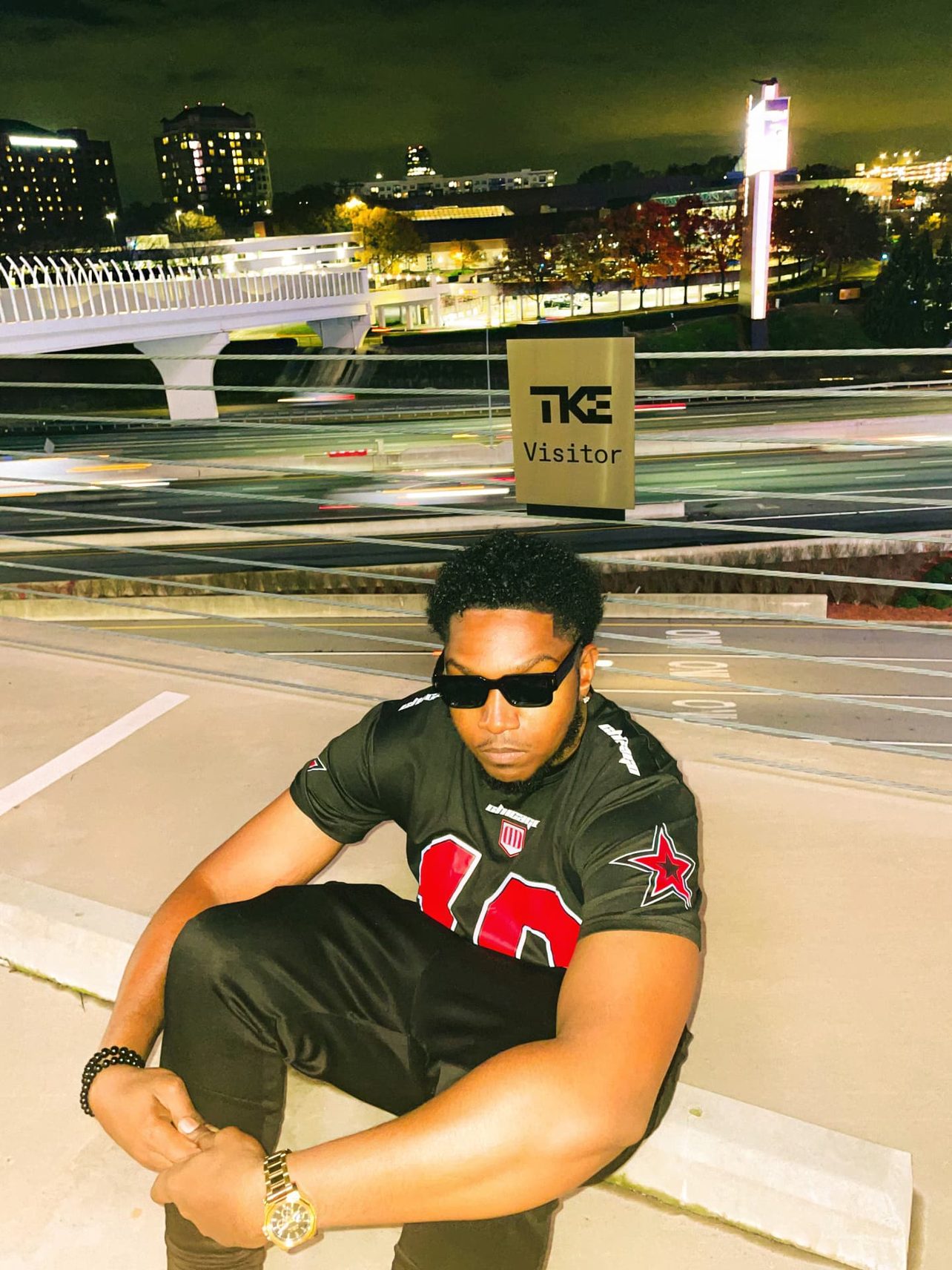 KAPOLLO seated on a parking deck at night in black sunglasses and a red-starred jersey, Atlanta skyline and highway light trails behind him.